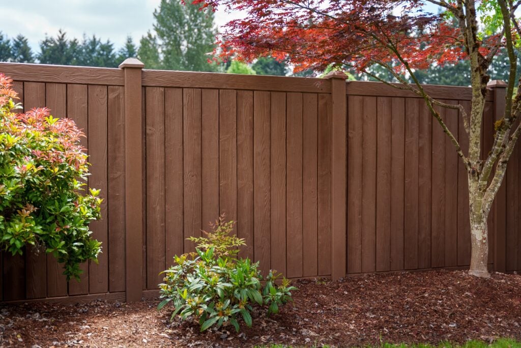 Brown privacy fence with vertical panels and wood grain texture, with shrubs and a small tree in front.