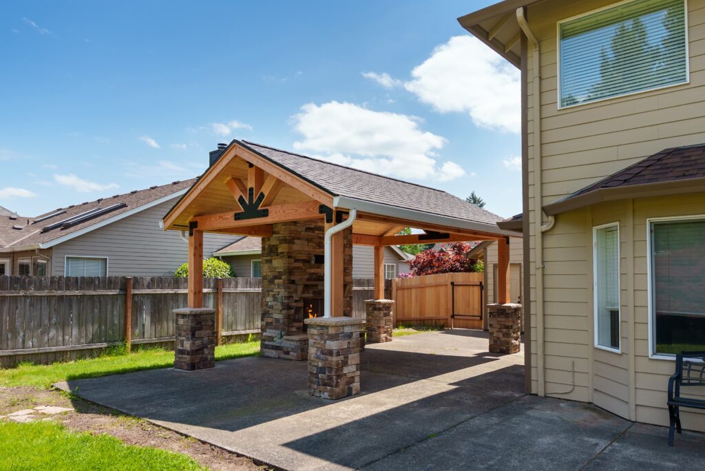 A covered outdoor patio featuring a stone fireplace under a timber frame roof, attached to the side of a house.
