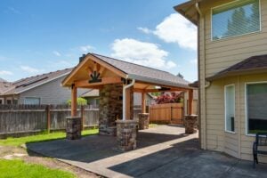 A covered outdoor patio featuring a stone fireplace under a timber frame roof, attached to the side of a house.