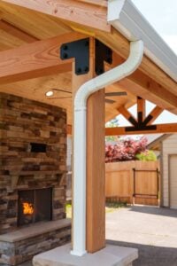 A white metal downspout attached to a wooden patio post, with an outdoor stone fireplace in the background.