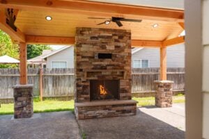 A stone outdoor fireplace with a fire burning inside, situated under a covered wooden patio structure.