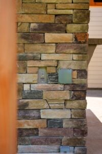 An outdoor stone wall featuring an integrated electrical outlet cover and a mounted doorbell device.