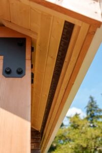 A close-up view of the underside of a wooden roof eave featuring a long, narrow ventilation strip, attached to a vertical timber post with a black metal bracket.