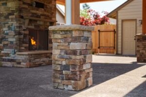A square stone patio pillar standing on a concrete surface, with an outdoor fireplace in the background.
