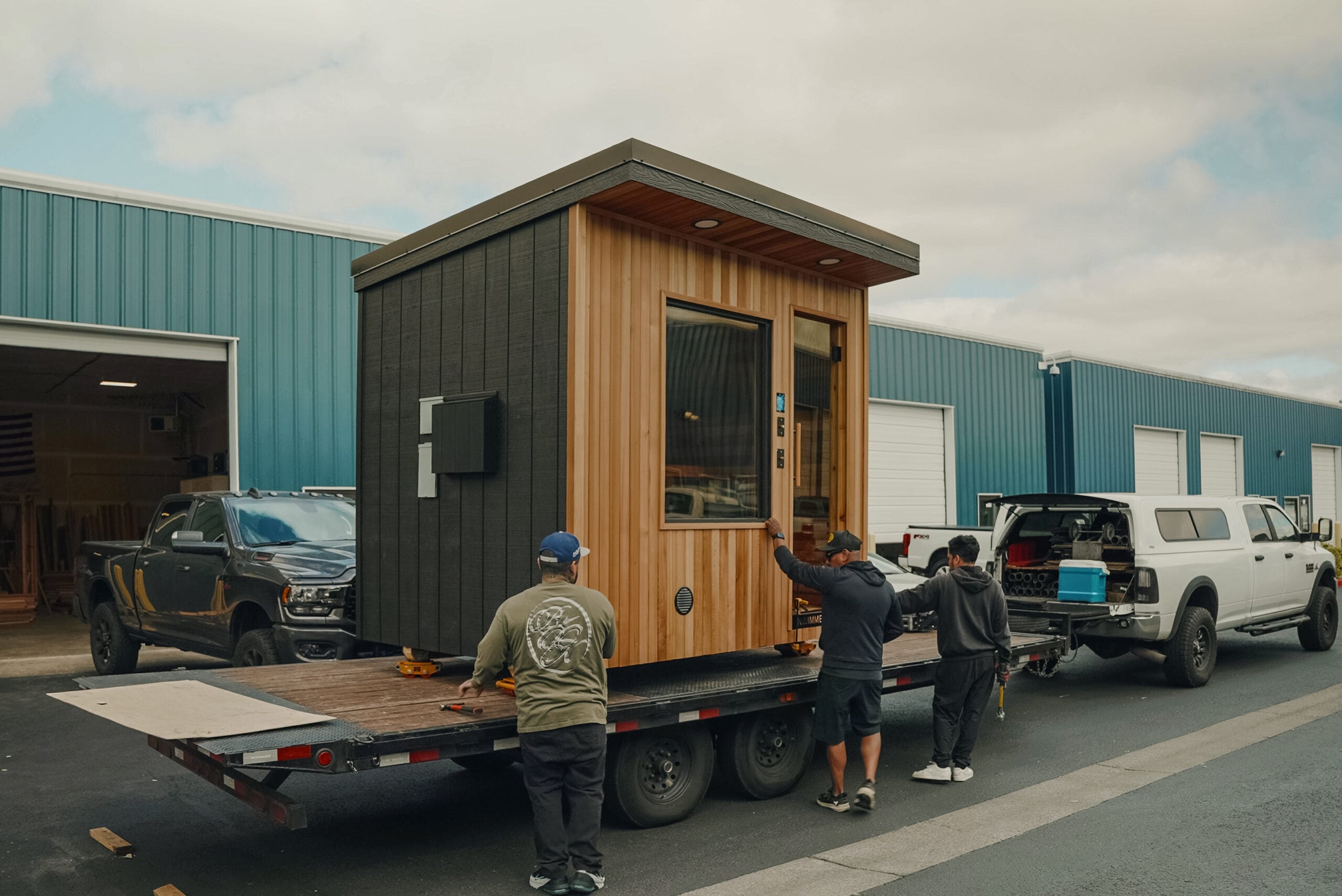 Three people load a small modern wooden tiny house onto a flatbed trailer, parked outside blue industrial buildings under a cloudy sky.