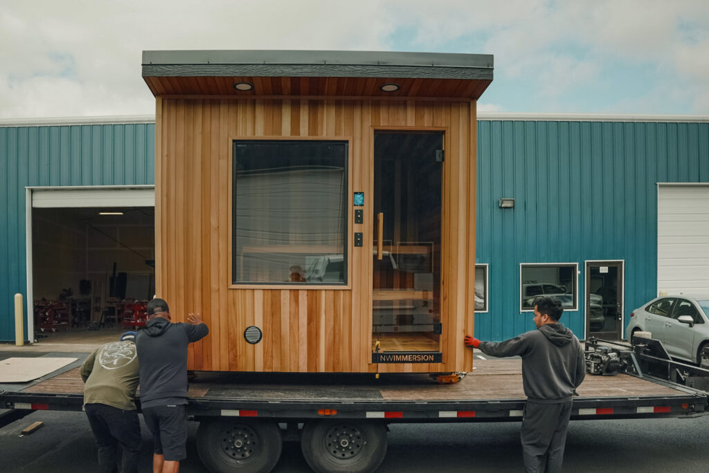 Three people move a small wooden cabin or sauna onto a flatbed trailer in front of a blue industrial building on a cloudy day.