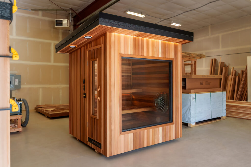 A wooden sauna with glass door and window sits inside a warehouse with unfinished walls and construction materials stacked nearby.