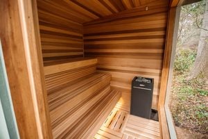 A modern wooden sauna with tiered benches, a sauna heater with stones, and a large window showing trees outside. The interior features smooth wood paneling and a warm, inviting atmosphere.