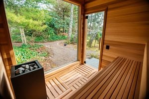 Interior of a wooden sauna with benches and a heater, featuring a large window and a glass door overlooking a green garden path surrounded by trees and plants.