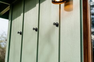 Close-up of a pale green exterior wall with three round black hooks and vertical trim. A portion of a lit outdoor lamp is visible at the top right corner.