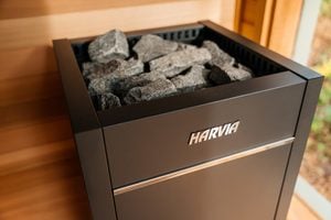 A black Harvia sauna heater filled with dark sauna stones, placed inside a wooden room with sunlight coming through a window.