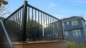 A close-up view of a black metal railing on a raised wooden deck with houses and trees in the background, and sunlight shining through the bars under a blue sky.