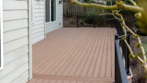 A newly built brown composite deck attached to a light gray house, with black metal railings and a small tree branch in the foreground. The yard is visible in the background.