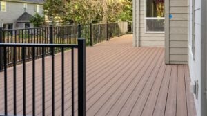 A spacious wooden deck with black metal railings is attached to a beige house. Trees and another house are visible in the background.
