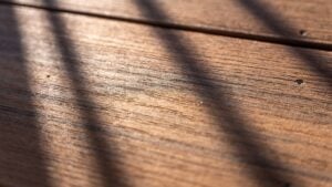 Close-up of wooden planks with a natural texture. Soft shadows, likely from railings or slats, fall diagonally across the sunlit surface. Two small screws or nails are visible on the right side of the planks.