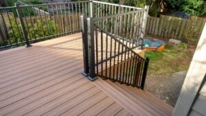 A wooden deck with brown planks features black metal railings and stairs leading down to a backyard with grass, trees, and a sandbox in the corner.