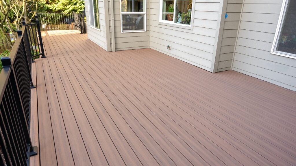 A spacious, clean wooden deck attached to a light-colored house with black railings and multiple windows, overlooking a leafy backyard.