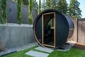 A cylindrical barrel sauna with a glass door in a backyard setting.