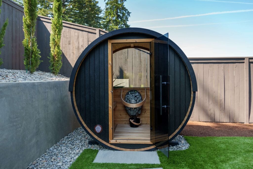 A black barrel sauna with a glass door in a backyard, featuring an interior heater with stones.