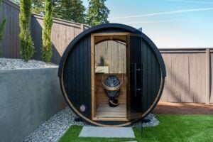 A black barrel sauna with a glass door in a backyard, featuring an interior heater with stones.