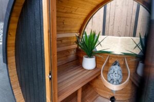 Interior view of a barrel sauna with a wooden bench, a potted plant, and a basket of sauna stones.