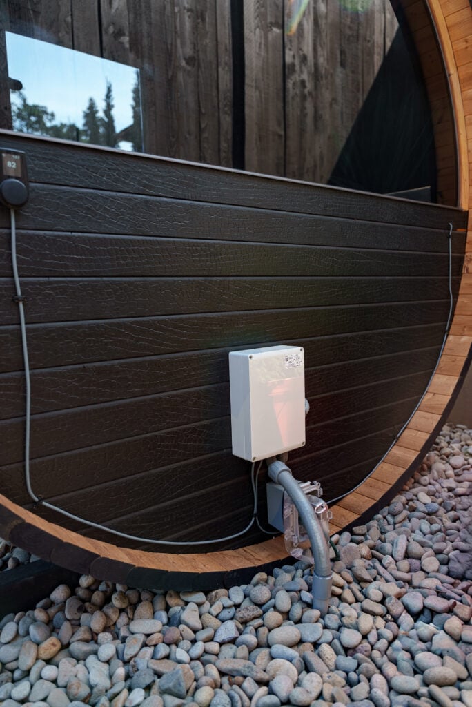 A white electrical box with cables and pipes is mounted on the side of a round, wooden outdoor structure. The ground is covered with small rocks.