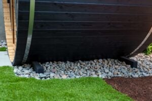 The base of a black barrel sauna resting on a bed of grey stones next to a patch of green grass.