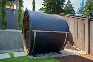 A black barrel sauna with a glass door sitting on a gravel patio in a backyard.