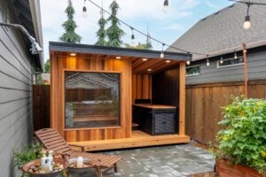 A cedar-paneled outdoor sauna with a window and hot tub on a stone patio, illuminated by string lights.