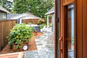 A view looking out from an open wooden door onto a backyard patio with a stone path, raised garden beds, and string lights.