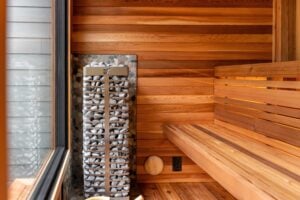 A sauna heater filled with stones standing next to a wooden bench in a cedar-paneled room.