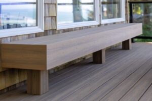 A long, rectangular wooden bench built into the side of a deck against a house with shingled walls and large windows.