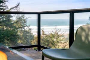 View of an ocean beach and trees from a wooden balcony with a glass railing and a green chair.