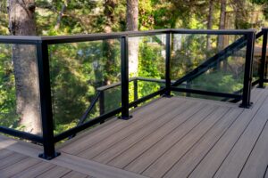 A deck with brown composite decking boards and a black metal railing with glass panels overlooking a wooded area.