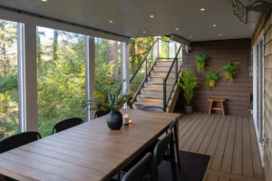 Covered outdoor dining area featuring a wooden table with a vase and candle, leading to a staircase and deck decorated with wall-mounted potted plants.