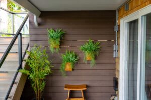 Three woven hanging baskets filled with ferns mounted on a brown horizontal-slatted wall, with a small potted bamboo tree and a wooden stool on the patio.