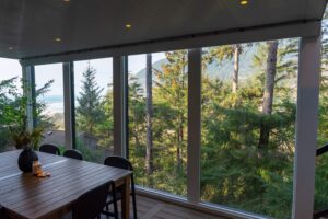 A dining table on a deck overlooking a forest and a distant ocean beach, viewed through large floor-to-ceiling windows.