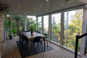 A dining table with six chairs on a covered deck overlooking a forest and distant water.