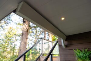 A view from under a porch roof showing a recessed ceiling light, a white support beam, a downspout, and a glass-paneled railing overlooking trees.