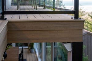 An elevated deck with wood-look composite planks and a black metal railing with glass panels overlooking a tree-lined view.