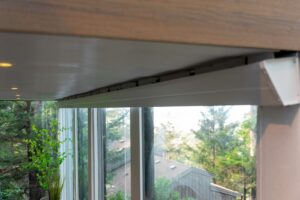 A white window blind casing mounted to the ceiling above a series of large windows overlooking a wooded area.