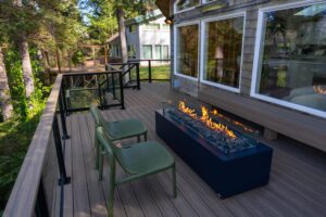 Two olive green chairs on a wooden deck next to a rectangular fire pit with a glass wind guard.