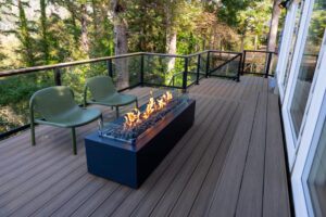 A rectangular outdoor fire pit with flames sits on a wooden deck next to two green chairs, with a forest view beyond the glass railing.