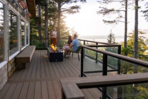 Three people sit and stand around a rectangular fire pit on a wooden deck overlooking a forest and the ocean at sunset.