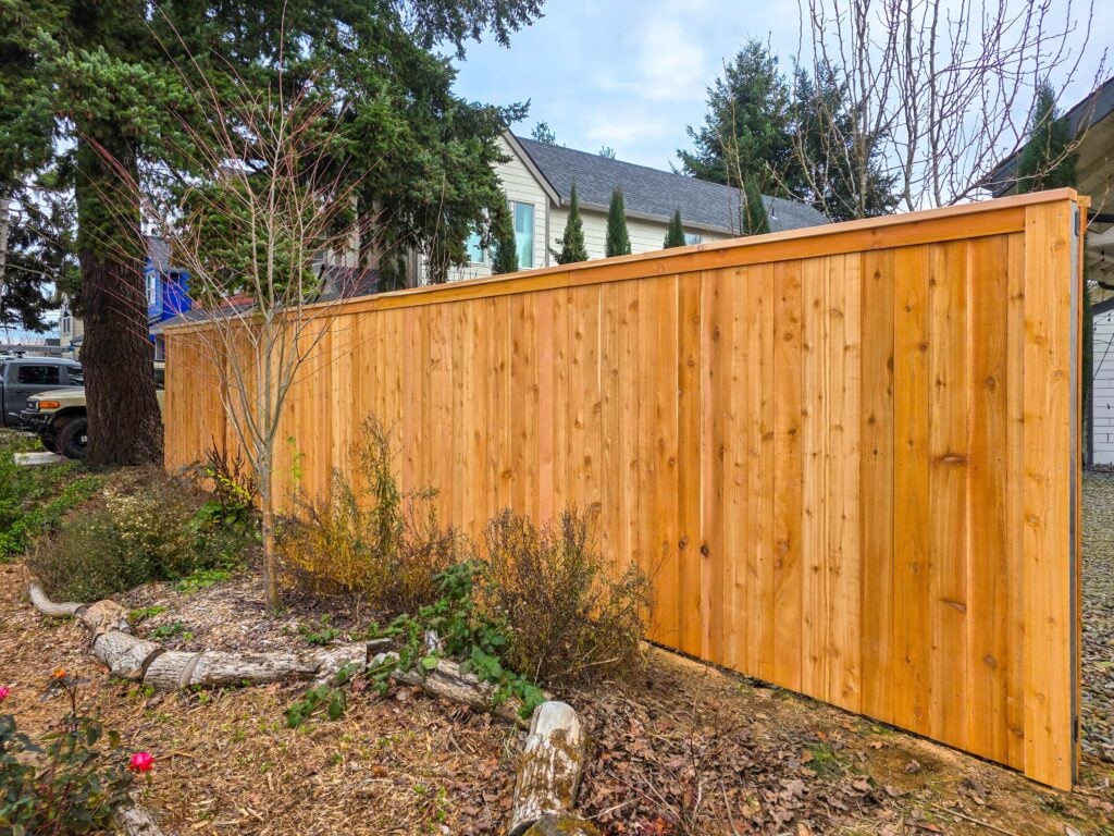 A tall, newly constructed wooden fence runs along a garden with mulch, shrubs, and a tree, separating the yard from a house and driveway in the background.