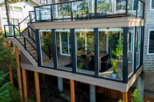A modern house with a wraparound glass-railed deck, an enclosed lower-level sunroom with a dining set, and an exterior staircase.