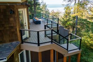 A multi-level outdoor deck featuring a rectangular fire pit and two chairs, overlooking a forest with a view of the ocean in the distance.