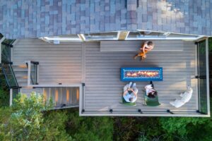 An aerial view of three people sitting on a deck around a rectangular fire pit with a dog standing nearby.
