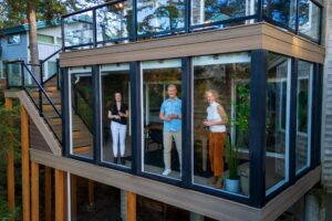 Three people holding wine glasses stand inside a modern sunroom with large windows on the second floor of a house.