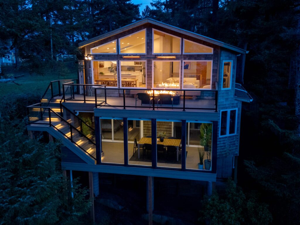 A two-story modern house at twilight with large glass windows, a fire pit on the upper deck, and illuminated interiors.